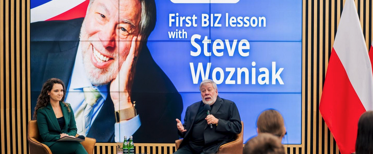 Z przodu sali pod wielkim ekranem z napisem First BIZ lesson with Steve Wozniak siedzi Steve Wozniak i mówi do trzymanego w ręku mikrofonu, obok niego kobieta w zielonym garniturze, przed nimi na sali siedzą ludzie.